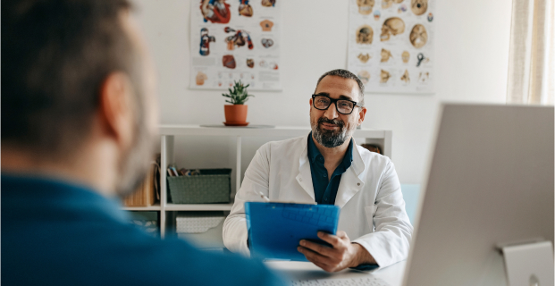 A doctor in a lab coat sits at a desk and speaks to a patient who is in the foreground.