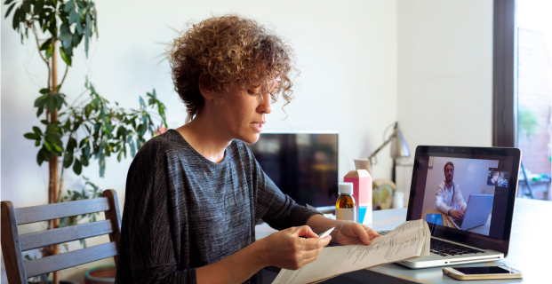 A woman sits at a desk as she consults with a telehealth specialist on her laptop.
