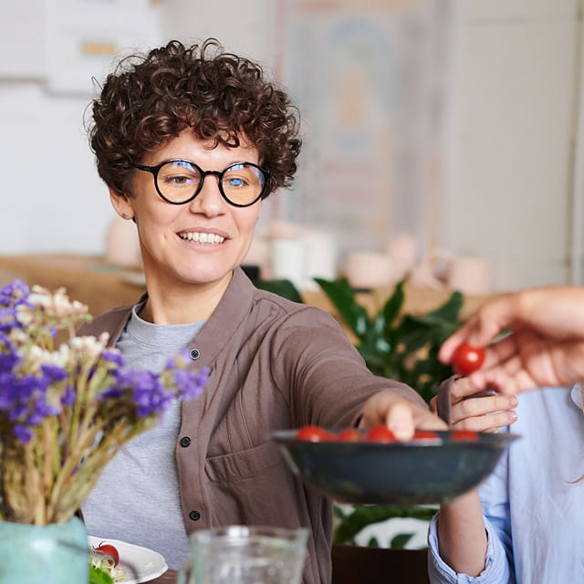 A woman with short, dark curly hair, wearing round glasses and holding a bowl of cherry tomatoes.
