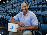 Albert Pujols sentado en las gradas de un estadio con una caja Cologuard en su regazo.